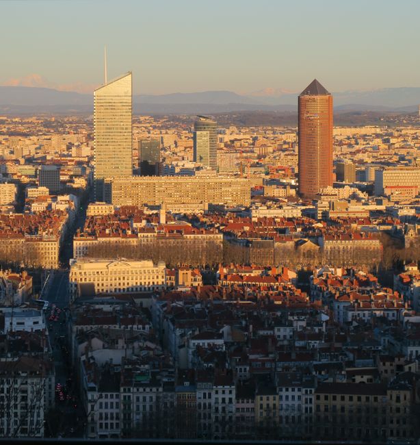 A panoramic view of Lyon's city skyline during daylight, featuring several high-rise office buildings including one with a distinctive sloped roof and another with a pointed spire, set behind an older