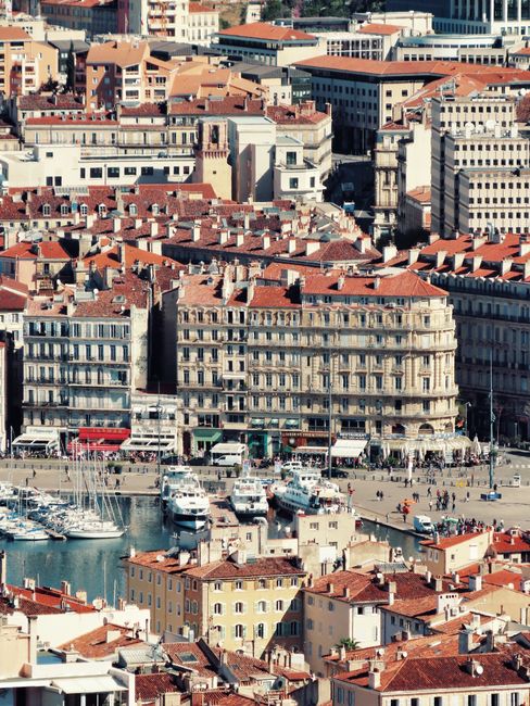 An aerial view of the historic Vieux Port area in Marseille, showing tightly packed buildings with red-tiled roofs, some featuring balconies with ornate railings. In the foreground, a marina is visibl