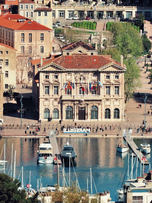 An aerial view of the historic Vieux Port area in Marseille, showing tightly packed buildings with red-tiled roofs, some featuring balconies with ornate railings. In the foreground, a marina is visibl