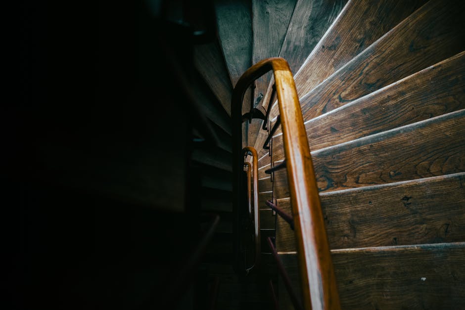 A straight flight of stone stairs inside a historic building, with a simple wooden handrail on the left side and an ornate black wrought iron railing on the right, leading up to an unseen upper level.